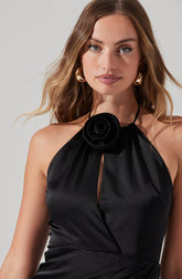 A woman is wearing a sleeveless black satin dress with a halter neckline, featuring a black fabric flower detail at the neck. The dress has a keyhole opening in the front. She accessorizes with large gold hoop earrings.